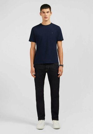 Chemise à manches courtes bleu marine en coton, associée à un jean foncé. Modèle se tenant debout avec des baskets blanches, les mains sur les côtés, fond neutre.
