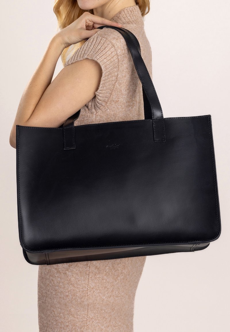 Femme vêtue d'une robe-pull beige sans manches tenant un grand sac cabas en cuir noir sur l'épaule, devant un fond uni.