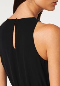 Kvinna i en ärmlös svart klänning med knäppning i nyckelhålet på baksidan av halsringningen, som visar nacke, axel och en del av ansiktsprofilen.