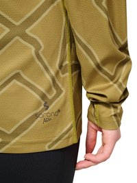 Close-up af person iført grøn mønstret langærmet top med "Sorona Agile" logo og sorte bukser, hånd afslappet ved siden af kroppen.