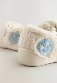 Chaussons crème confortables avec une surface en polaire texturée, ornés d'un motif de visage souriant bleu et d'une semelle en caoutchouc de couleur claire.