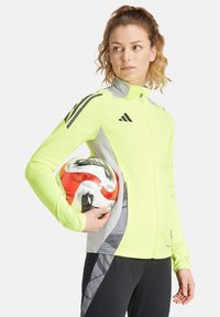 Veste haute visibilité de couleur jaune fluo avec des accents gris, fermeture éclair à l'avant et coupe ajustée, tenue par une personne avec un ballon de football à motifs.