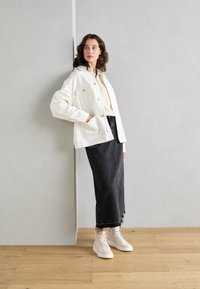 Weißes oversized Jackett mit großen Knöpfen, getragen über einem hellen Oberteil und einem schwarzen Denim-Rock. Das Model trägt weiße High-Top-Sneaker.