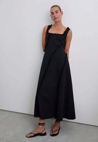 Zwarte maxi-jurk met vierkante halslijn en gerafelde randen, met een relaxte pasvorm en diagonale naaddetails, gecombineerd met sandalen met banden.
