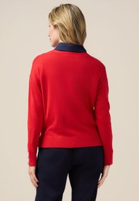 Pull en tricot rouge avec un col marine, manches longues et accents de boutons aux poignets. La coupe est décontractée, avec un ourlet droit.