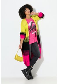 Langer gestrickter Cardigan in Gelb, Pink und Schwarz, getragen über einem pinken Grafik-T-Shirt, kombiniert mit dunklen Jeans und schwarzen Kampfstiefeln. Kleine gelbe Handtasche.