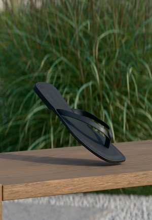 Sandale noire de type tongs équilibrée sur son talon sur un banc en bois avec de hautes herbes vertes floues en arrière-plan.