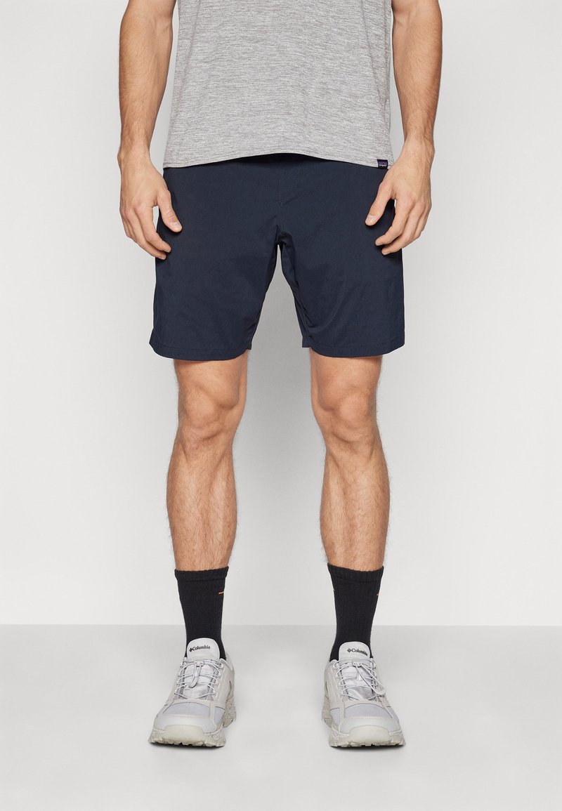 Marineblå shorts med en glatt tekstur, midt på låret lengde, og en svak glans. Brukes med en grå t-skjorte og grå joggesko. Svarte sokker synlige.