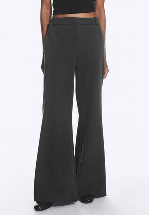 Personne portant un pantalon large taille haute gris foncé associé à un top court noir, debout avec les mains détendues sur les côtés.