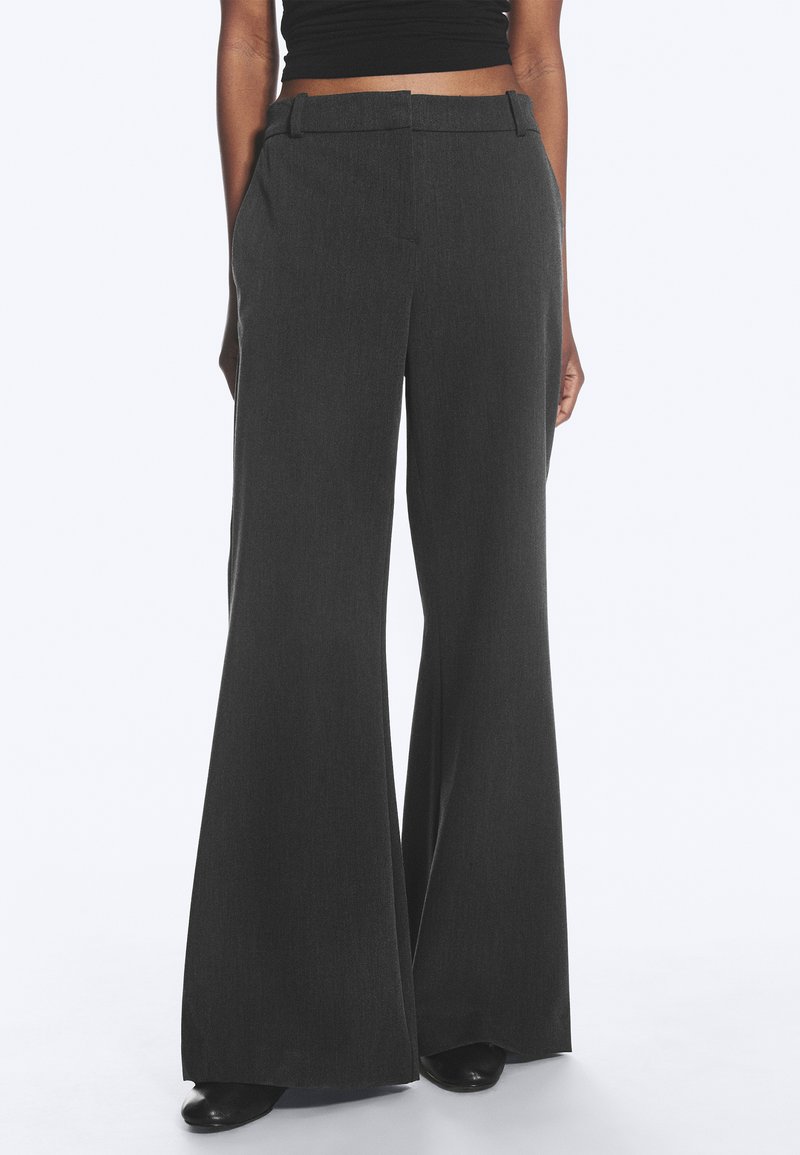 Personne portant un pantalon large taille haute gris foncé associé à un top court noir, debout avec les mains détendues sur les côtés.