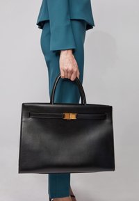 Svart läderhandväska med en strukturerad design, guldaccentdetaljer och ett handtag upptill, hålls av en person i en teal-outfit.