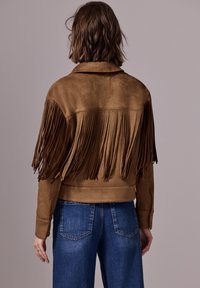 Brun mockajacka med fransar på baksidan, kort design och skjortkrage. Kombinerad med blå denimjeans.