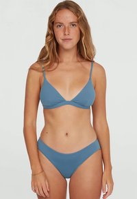 Femme aux longs cheveux ondulés portant un bikini deux pièces bleu, se tenant devant un fond gris clair uni.