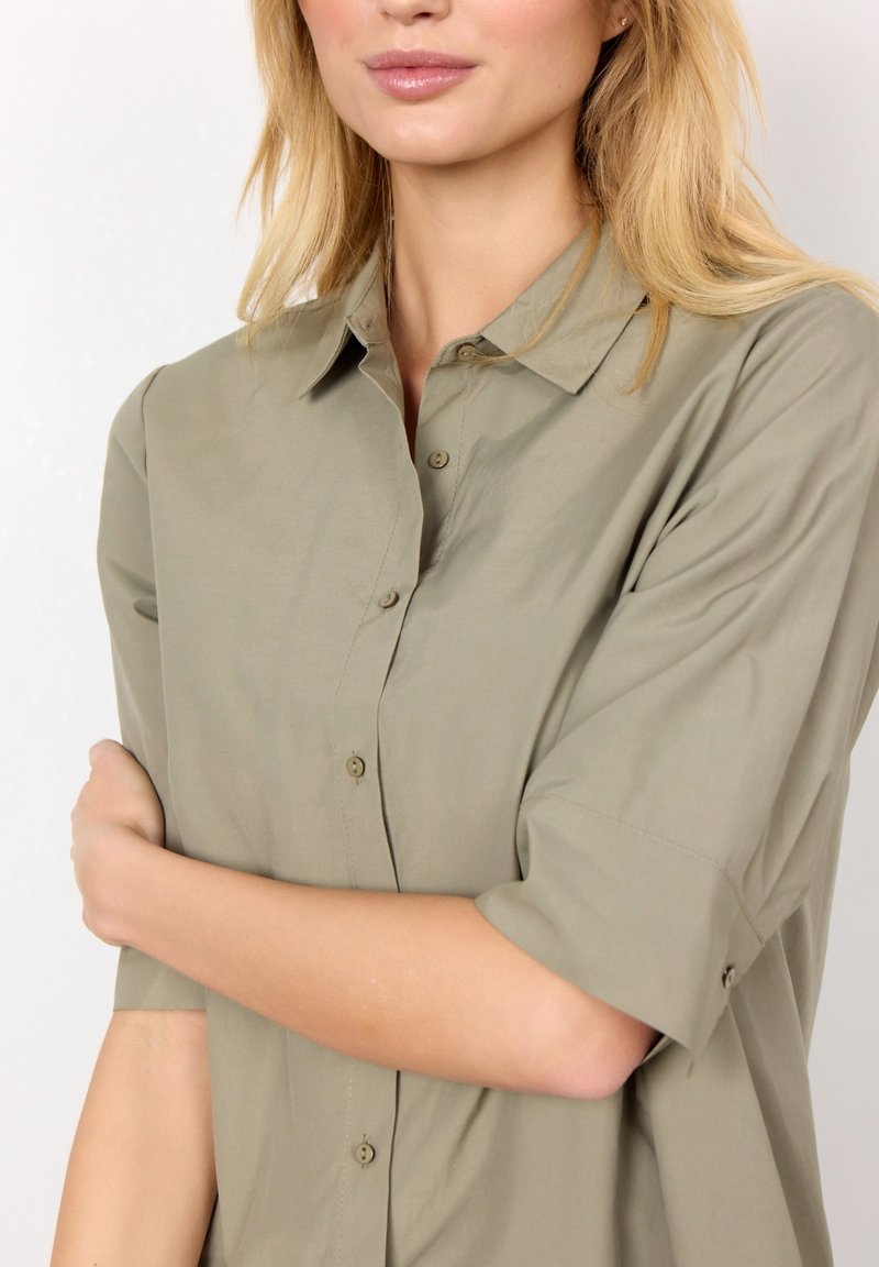 Camicia khaki con bottoni realizzata in tessuto leggero, con colletto, maniche corte e vestibilità rilassata. Texture liscia, dettagli minimi.