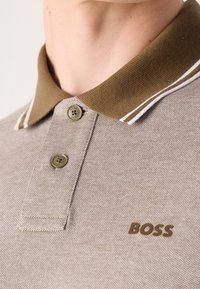 Polo beige avec un col tricoté marron, featuring des rayures blanches, deux boutons marron, et le logo "BOSS" en marron sur la poitrine.