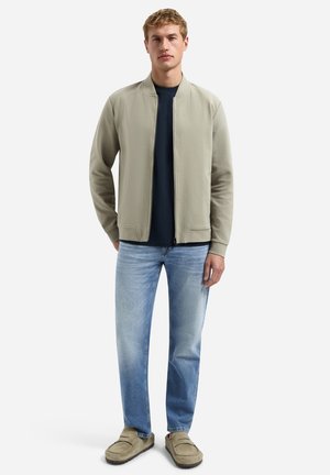 Jeune homme debout, portant une veste zippée beige, une chemise bleu marine, un jean bleu clair et des chaussures beiges sans lacets, sur fond blanc.