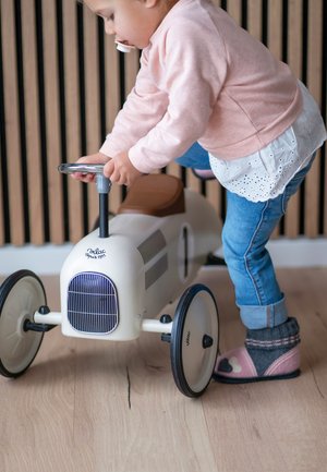 Crème-kleurige loopauto met een bruine zitting, chrome stuur en zwarte wielen. Kind dat met de auto op een houten vloer speelt.