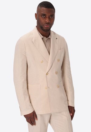 Uomo con capelli corti che indossa un blazer beige doppiopetto e pantaloni coordinati su uno sfondo bianco uniforme.