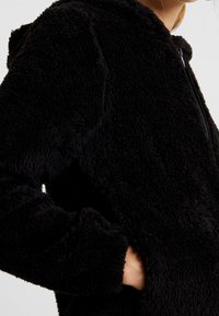 Svart fleecehoodie med en mjuk, texturerad yta, dragkedja och sidfickor. Har en huva och långa ärmar för värme.