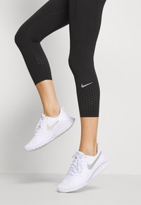Czarne legginsy capri z perforowanym detalem, połączone z białymi butami do biegania Nike z odblaskowym srebrnym swooshem i teksturowanym materiałem.
