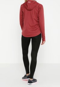 Maglione rosso a maniche lunghe con cappuccio dalla consistenza liscia, abbinato a leggings neri con una vestibilità adatta e cerniere alla caviglia. Scarpe da ginnastica nere.