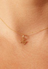 Collier en or avec un pendentif stylisé en forme de lettre "M" proéminente. La chaîne est délicate et brillante, posée sur un fond couleur peau.