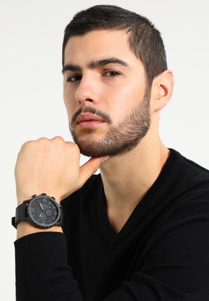 Uomo barbuto che indossa un maglione nero a scollo a V, appoggiando il mento sul pugno, mostrando un orologio da polso nero con grandi numeri.