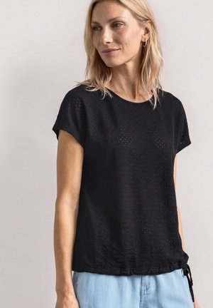 Femme aux cheveux blonds portant un haut noir texturé à manches courtes et un pantalon bleu clair, regardant légèrement vers sa gauche sur un fond uni.