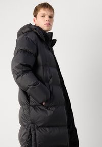 Sort pufferjakke med hætte, der har en quiltet design, lynlås lukning og sidelommer. Materialet ser skinnede og isoleret ud.