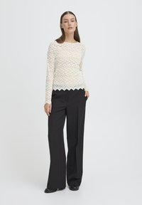 Lace langærmet top i creme med bølget kant, parret med højtaljede sorte bukser, skaber en lagdelt teksturel effekt.