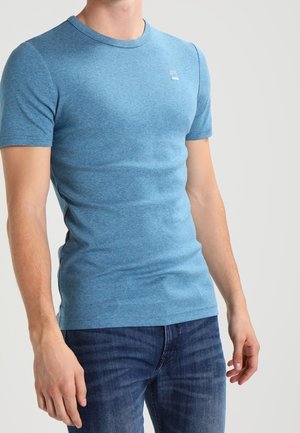 Mann trägt ein eng anliegendes blaues T-Shirt mit kurzen Ärmeln und dunkelblaue Jeans, steht vor schlichtem hellem Hintergrund.