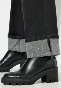 Botines negros hasta el tobillo con suela gruesa, hechos de cuero brillante. Los vaqueros tienen un puño gris claro con un sutil detalle brillante.