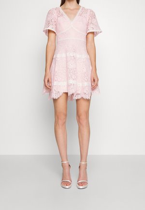 Femme portant une robe courte en dentelle rose avec un col en V et des manches volantes, associée à des sandales blanches à talons hauts avec bride à la cheville, debout sur un sol gris.