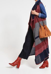 Person som bär röda ankelstövlar, mörka avklippta jeans, blå skjorta, lång randig väst, bär en beige mocka axelväska, går mot en vit bakgrund.