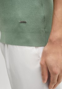 Pullover verde a maglia texturizzata con orlo a coste e accento del logo argento "JOOP!"; indossato con pantaloni bianchi, evidenziando un design aderente.