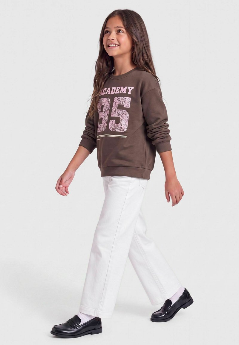 Jeune fille marchant, portant un sweat-shirt marron avec un pantalon blanc, des chaussettes blanches et des mocassins noirs, souriante et regardant vers le haut.