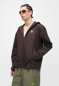 Καφέ ελαφρύ μπουφάν με φερμουάρ και κουκούλα, με λευκό λογότυπο Adidas, φορεμένο πάνω από λευκό αμάνικο τοπ και συνδυασμένο με χακί παντελόνι.