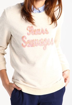Sweatshirt couleur crème avec des lettres texturées roses et oranges portant l'inscription "Fleurs Sauvages". Col rond et poignets côtelés ; tissu doux et décontracté.