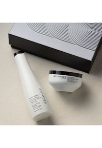 Deux bouteilles blanches de shampooing et de traitement sont placées sur une surface beige clair, avec une boîte grise texturée au motif tourbillonnant en arrière-plan.