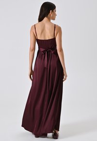 Bordeaux satijnen maxi-jurk met dunne banden, een ingesnoerd middel en een strikdetail op de rug, met een vloeiende silhouet en een zachte textuur.