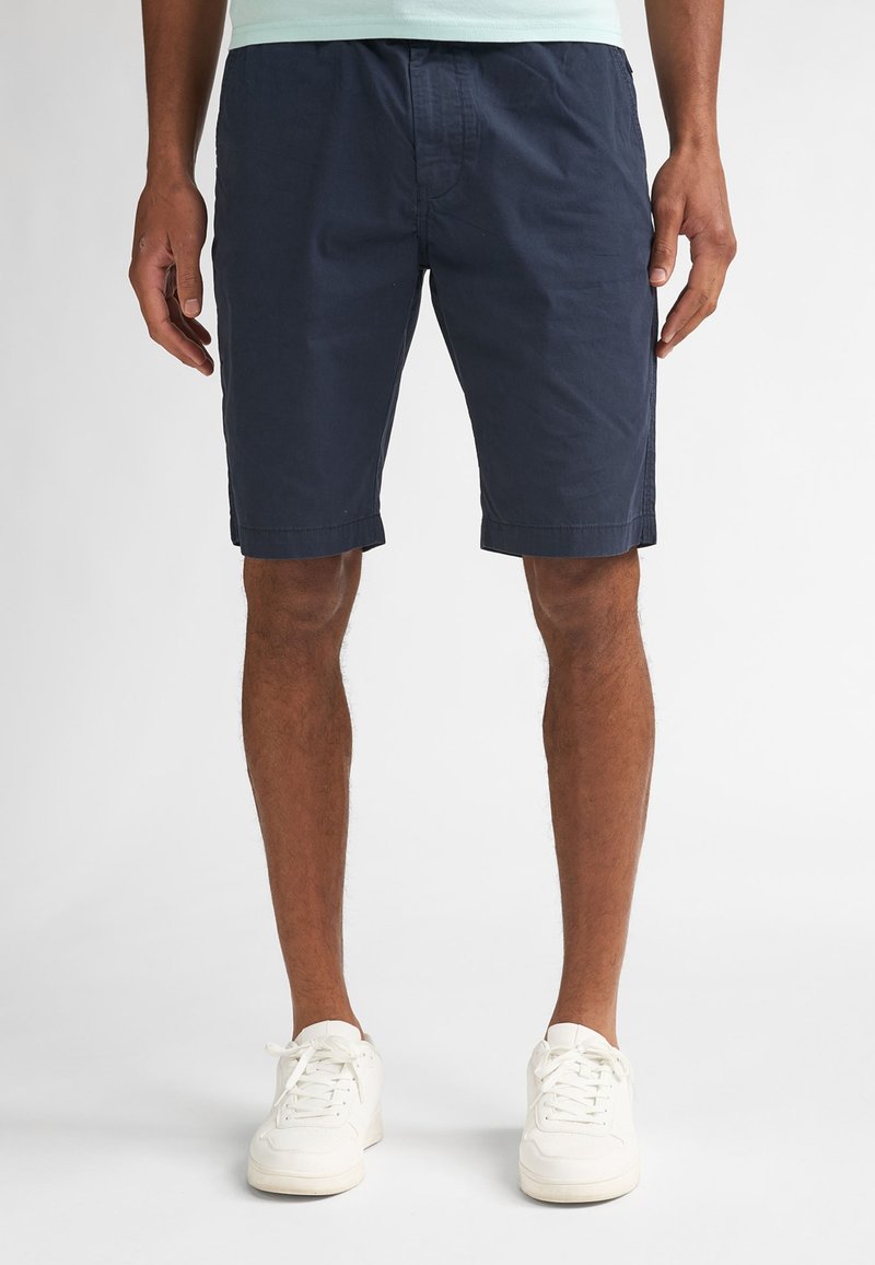 Hombre con pantalones cortos azul marino, zapatillas blancas y una camisa de color claro, de pie contra un fondo blanco liso.