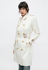 Fehér, kétsoros arany gombos ballonkabát, derékrésznél övvel, állítható mandzsettákkal. Sima anyag, klasszikus gallér kialakítás, oldalzsebek.