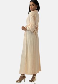 Maxikjole i blødt, struktureret ferskenstof med bælte i taljen og lange ærmer. Har sideslidser og er stylet med lyse hæle.