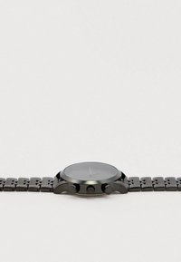 Crni metalni ručni sat s povezanim narukvicom i tri bočne tipke na svijetlosivoj pozadini, prikazan iz razine brojčanika.