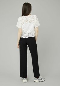 Blouse en dentelle blanche avec des motifs floraux découpés, associée à un pantalon large noir et des baskets argentées, vue de dos.