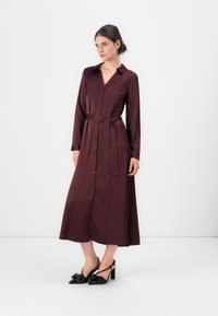 Burgundi hosszú ujjú ingruha gombos elülső résszel, övvel a derekán és sima textúrával. A modellen fekete, masnival díszített slingback magassarkú cipők vannak.