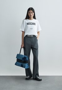 Λευκό μπλουζάκι Moschino με μαύρο τύπωμα, γκρι παντελόνι με ψηλή μέση, μπλε τσάντα από τζιν, μαύρα αθλητικά παπούτσια με άσπρες σόλες, στέκεται πάνω σε γκρι φόντο.