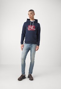 Felpa con cappuccio blu navy con stringhe bianche e stampa grafica rossa. Indossata con jeans azzurri strappati e sneaker marroni.