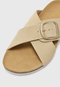 Sandalo in suede beige con cinturini incrociati, dotato di una fibbia rettangolare in argento. Sottopiede marrone chiaro e suola in gomma bianca.