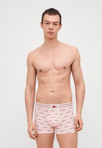 Heren boxershorts van lichtroze katoen met een patroon van het LACOSTE logo over de hele stof. Voorzien van een contrasterende tailleband met rode strepen. Fitted ontwerp.